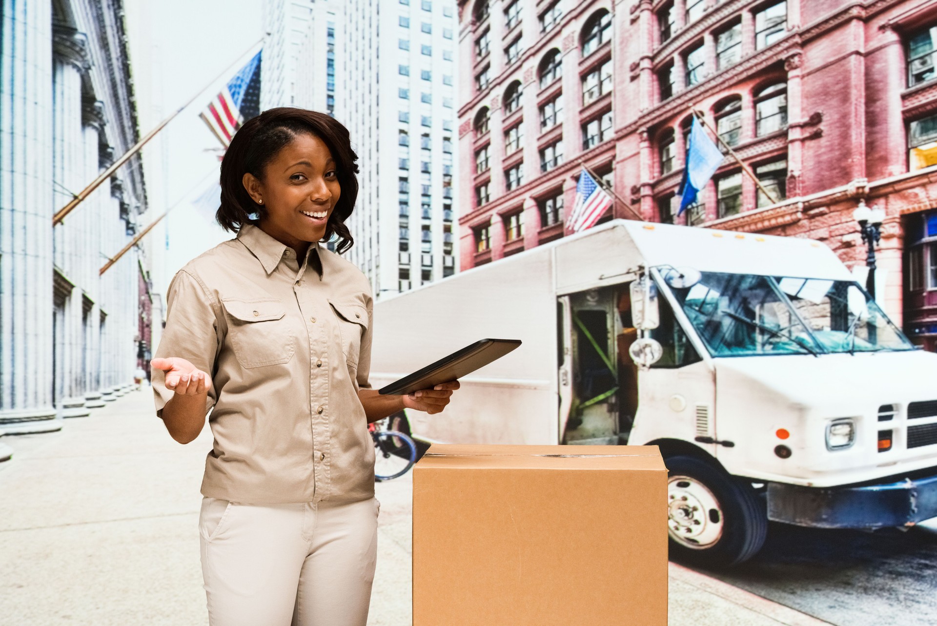 African-american ethnicity female delivery person standing at the delivery van wearing button down shirt and holding cardboard and using digital tablet